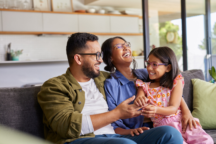 Parents playing with daughter at home Cheerful middle eastern family with eyeglasses of three having fun together at home. Mixed race parents talking with their little girl in living room. Cheerful family with specs embracing and playing with beautiful indian daughter girl in arms.